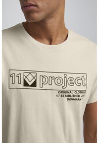 Beige bomull t-skjorte med rund hals og svart grafisk trykk som leser "11 prosjekt," "original klær," og "etablert 2020 Danmark."