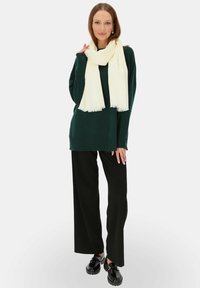 Ciemnozielony sweter z dzianiny, czarne spodnie szerokie w nogawkach oraz długi biały szalik z frędzlami na końcach. Czarne buty dopełniają całość.