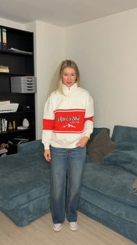 Vrouw die binnen staat, draagt een witte en rode sweatshirt met de tekst "Après Ski with me", een blauwe spijkerbroek en witte sneakers, naast een blauwe bank en een zwarte plank.