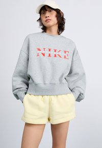 Sweatshirt gris avec logo "NIKE" rouge, poignets côtelés, porté avec un short jaune avec un swoosh Nike blanc. La casquette est de couleur crème.