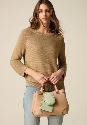 Giovane donna con maglione beige e jeans chiari che tiene una borsa color cuoio con una piccola pochette verde attaccata su sfondo semplice.