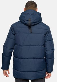 Marineblå polstret jakke med hætte, der har vandret syninger og en sort remdetalje på ryggen, lavet af glat, let materiale.