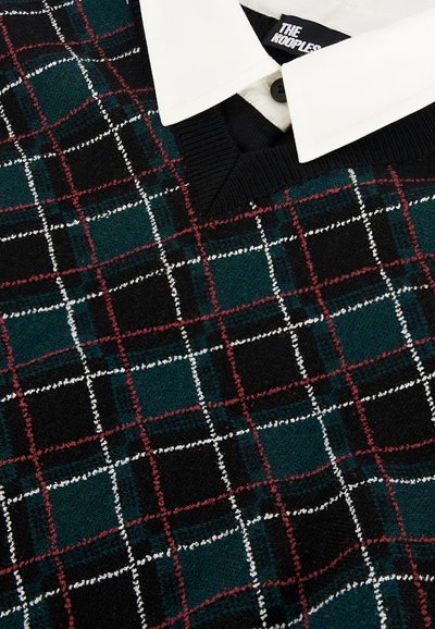 Sweater de malha em padrão xadrez verde, preto e vermelho, com linhas brancas e vermelhas, combinado com uma camisa branca de colarinho por baixo.