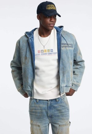 Veste en denim avec une finition bleu délavé, fermeture éclair à l'avant et capuche, superposée sur un sweatshirt graphique blanc. Porte avec un pantalon bleu clair assorti.