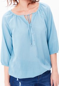 Blusa azzurro chiaro con maniche a tre quarti, scollatura a girocollo, dettaglio con fiocco frontale e patta con bottoni. Tessuto morbido con una sottile texture.
