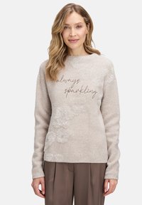 Jasno beżowy sweter z miękkiej tkaniny, z błyszczącymi włóknami i haftowanym wzorem "zawsze błyszczący" z przodu.