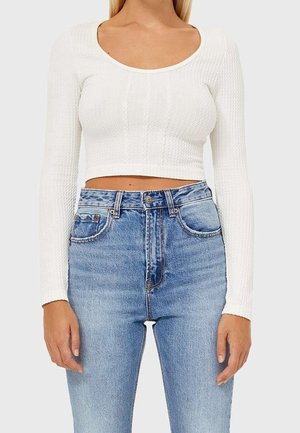 Femme portant un pull court à manches longues blanc texturé et un jean en denim bleu clair taille haute, sur un fond uni.
