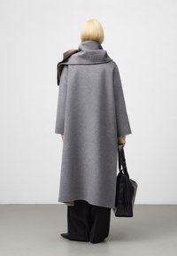 Grå ullrock med hög krage och lös passform, kombinerad med svarta byxor. En svart väska hålls av modellen och visar upp en minimalistisk design.