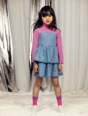Young girl standing wearing a pink-striped turtleneck, blue polka-dot dress, pink socks with blue stripes, and white sneakers.