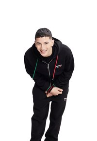 Felpa con zip nera con accenti verdi e rossi, abbinata a pantaloni della tuta neri. Presenta il logo sul petto e sulla gamba sinistra dei pantaloni. Tessuto morbido al tatto.