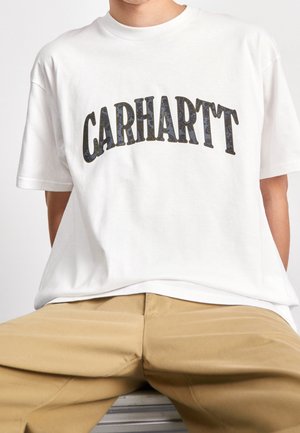 Person iført en hvid Carhartt t-shirt med buet sort mønstret logo, kombineret med beige bukser, siddende mod en ensfarvet baggrund.