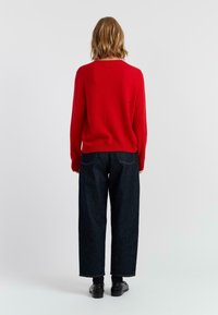 Roter, gestrickter Pullover mit Rundhalsausschnitt, kombiniert mit dunkelblauen, weiten Jeans und schwarzen Schuhen. Glatte Textur und schlichtes Design.