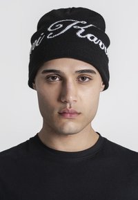 Zwarte gebreide beanie met witte geborduurde tekst. Glad materiaal en nauwsluitende pasvorm. Draagt een persoon in een zwart shirt tegen een effen achtergrond.