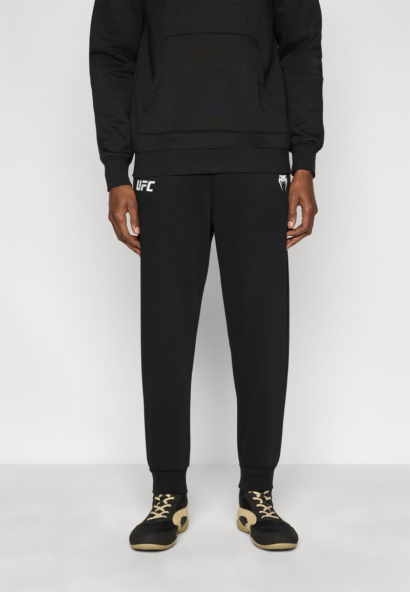 Μαύρο φούτερ με μπροστινή τσέπη, συνδυασμένο με αντίστοιχο μαύρο παντελόνι φόρμας με λευκό λογότυπο UFC. Φορεμένο με μαύρα και χρυσά αθλητικά παπούτσια.