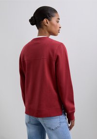 Sudadera roja con cuello redondo y puños acanalados, con una costura horizontal en la parte trasera. Se lleva con vaqueros azul claro.
