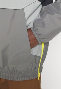 Rompevientos gris que presenta una combinación de tejido texturizado y liso, puños elásticos y un acento de cremallera en amarillo neón en el bolsillo delantero.
