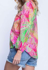 Värikäs pusero, jossa on vaaleanpunainen, vihreä ja keltainen trooppinen lehtikuvio. Siinä on pitkät hihat ja väljähkö istuvuus, yhdistettynä farkkushortseihin.