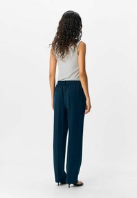 Femme aux cheveux bouclés se tient de dos, portant un top sans manches gris, un pantalon large bleu marine et des sandales à talons noires.