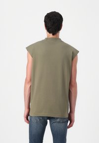 Camiseta sin mangas de color verde oliva fabricada en algodón, con un corte holgado y un cuello redondo, combinada con jeans azules. Diseño simple sin patrones.