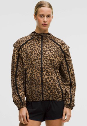 Žena v bunde so zipsom s leopardím vzorom a čiernymi pruhmi, v kombinácii s čiernymi krátkymi nohavicami, stojaca na jednoduchom pozadí.