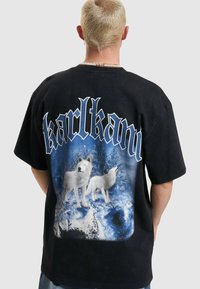 Svart bomull T-skjorte med et stort grafisk motiv av hvite ulver mot en blå fjellbakgrunn, med teksten "karlkani" i fet skrift.