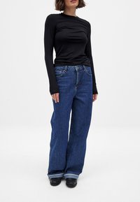 Svart långärmad skjorta i kombination med löst sittande, mörkblå jeans. Jeansen har knapp- och dragkedjestängning, med synliga sömmar och uppvikta mudd.