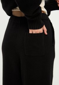 Černé pletené kalhoty s kulatým pasem mají na boku čtvercovou kapsu. Tkanina vypadá měkce a mírně texturovaně.