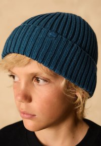 Temno zeleno modra rebrasta pletena kapa z majhnim emblemom na strani, ki jo nosi otrok s kratkimi blond lasmi na nevtralni ozadju.