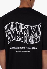 Zwarte katoenen T-shirt met gedurfde, vervormde witte tekst: "DROPSIZE" en "DROPSIZE CLUB / EST. 2020" op de achterkant. Casual fit.