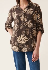 Chemise boutonnée marron avec un motif de feuilles tropicales en crème. Manches courtes à revers, coupe décontractée, fabriquée en tissu léger.