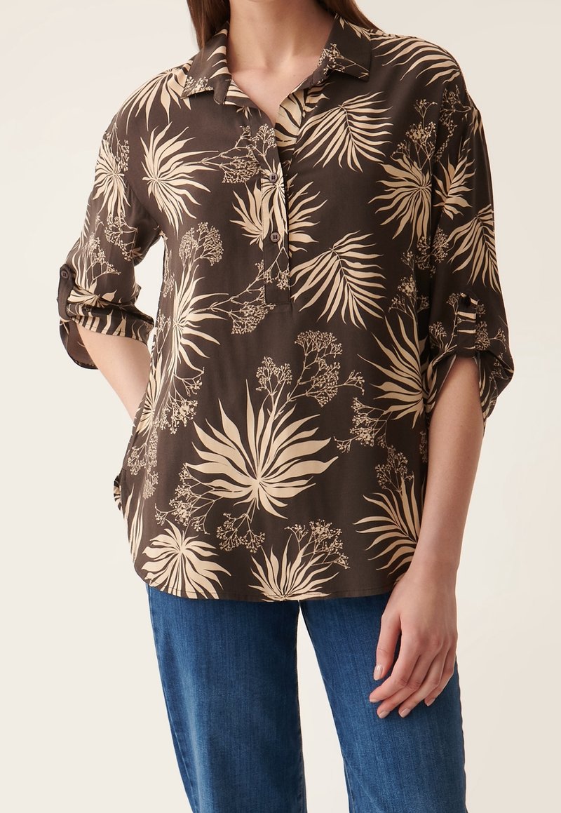 Chemise boutonnée marron avec un motif de feuilles tropicales en crème. Manches courtes à revers, coupe décontractée, fabriquée en tissu léger.