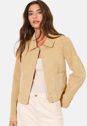 Woman with long brown hair wearing a beige suede jacket over a light pink top and light-colored pants, posing with a neutral expression.