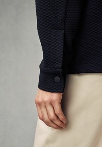 Sweatshirt bleu marine texturé avec un motif en diamant et un poignet boutonné. La main repose sur un tissu clair, montrant des matériaux contrastants.