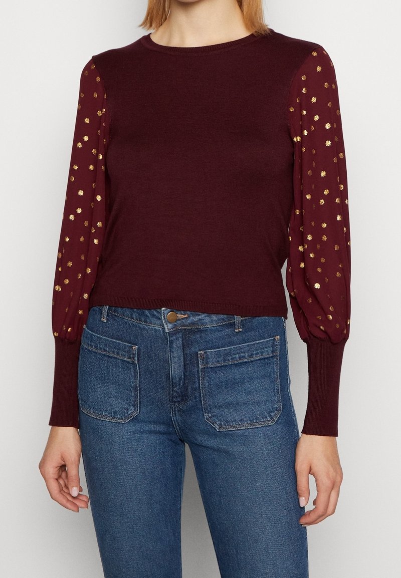 Pull bordeaux avec des manches ballon en voile ornées de pois dorés, associé à un jean bleu taille haute avec des poches plaquées à l'avant.
