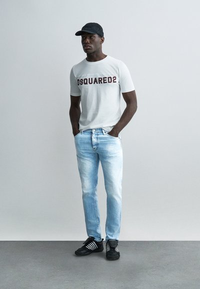 Homme portant un t-shirt blanc DSQUARED2, un jean bleu clair, une casquette noire et des baskets noires, debout les mains dans les poches devant un fond gris.