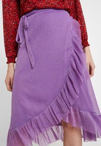 Fialová zavinovací sukně s texturovanou látkou, zdobená volánkovým lemem a páskem v pase, který zvýrazňuje její vrstvený design.