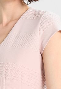 Blush-pink strukturierte Bluse mit kurzen Ärmeln, die horizontale erhabene Muster und einen V-Ausschnitt aufweist. Glatter Stoff mit subtilen Details.