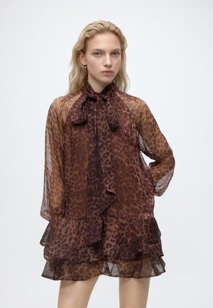 Jeune femme aux cheveux blonds ondulés portant une robe marron transparente à imprimé léopard, avec un col noué et un ourlet volanté.