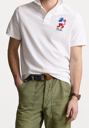 Homme portant un polo blanc avec un logo lion rouge et bleu et le texte « Polo », pantalon vert, ceinture rayée et montre marron, mains dans les poches.