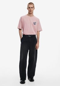 Light pink cotton t-shirt with a chest graphic, paired with loose-fitting black denim pants and black shoes. Simple design and relaxed fit.