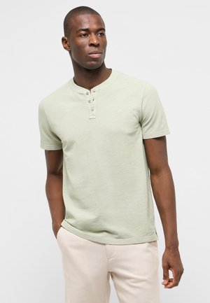 Mann trägt hellgrünes kurzärmliges Henley-Shirt und beige Hose, steht mit einer Hand in der Tasche und schaut seitlich vor einem schlichten Hintergrund.