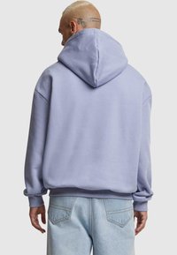 Felpa con cappuccio viola chiaro, dalla vestibilità rilassata, con una consistenza morbida e polsini a coste. Indossata con jeans in denim azzurro chiaro con tasche posteriori.