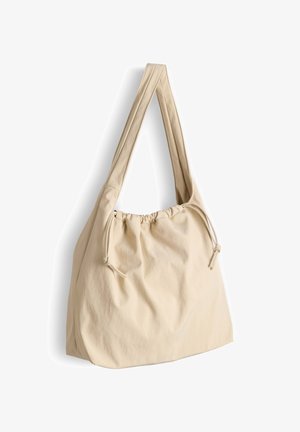 Borsa a spalla in tessuto beige con ampie cinghie e chiusura a coulisse, texture morbida, appesa su uno sfondo bianco.