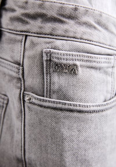 Gray denim jeans featuring a back pocket with a metal E.A logo and a small snap button. The fabric has a textured finish.