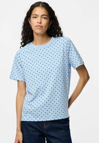 Camiseta de manga corta azul clara con lunares negros. Cuello redondo, corte relajado, tejido suave y diseño sencillo. Combinada con jeans oscuros.