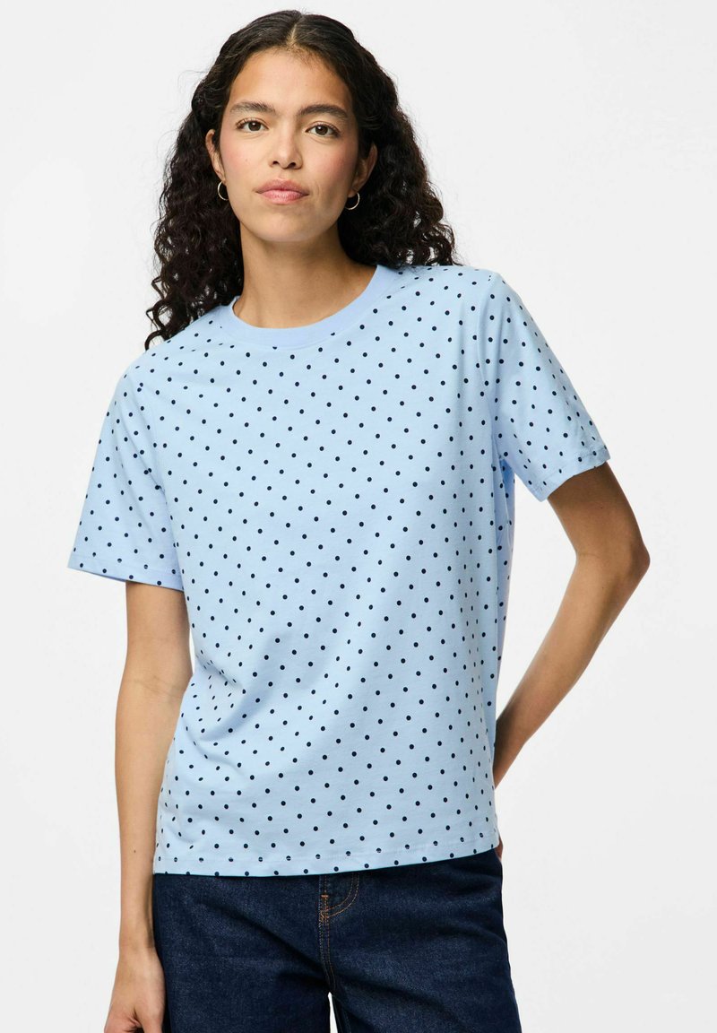 Camiseta de manga corta azul clara con lunares negros. Cuello redondo, corte relajado, tejido suave y diseño sencillo. Combinada con jeans oscuros.