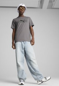 Grå kortærmet skjorte med sort logo, lyseblå vide jeans, hvid bucket hat og sporty sorte og hvide sneakers.
