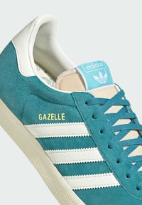 Tīruma zamšādas sporta apavi tumši zaļā krāsā ar baltām ādas akcentiem, ar trim baltām sliedēm un zelta "GAZELLE" logo. Gaiši zila mēle un teksturēta zole.