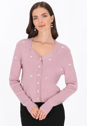 Femme portant un cardigan violet clair avec des boutons en perle et de petites décorations florales blanches, associé à un pantalon noir.