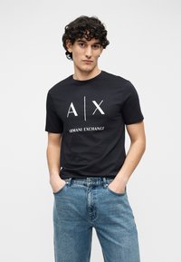 Μαύρο βαμβακερό t-shirt με το λευκό λογότυπο "A|X" και το κείμενο "ARMANI EXCHANGE". Συνδυασμένο με ανοιχτό μπλε τζιν παντελόνι, σε άνετη γραμμή.